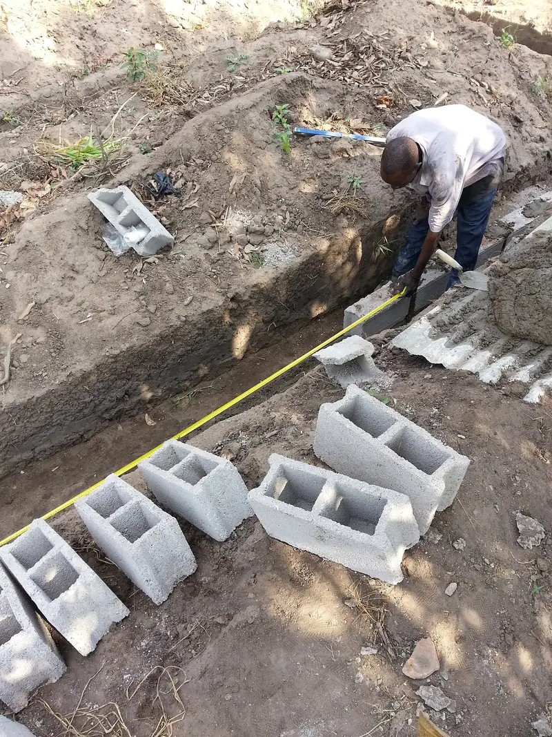 Maçon qui mesure une semelle de fondation, blocs de béton alignés au bord de la fouille