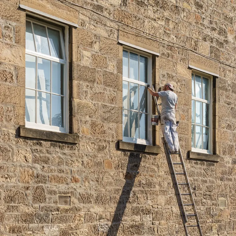 Peintre sur une échelle qui reprend les fenêtres d’une façade en pierre